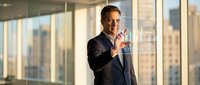 Professional businessman in dark suit pointing forward in modern office with city view through large windows during golden hour lighting.