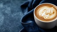 Hot cappuccino with artistic leaf latte art pattern in white ceramic cup on dark blue textured background with drapery and copy space for cafe menu.