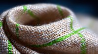 Natural burlap fabric with green thread pattern in macro shot showing detailed textile weave structure and organic fiber texture against dark background.