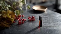 Small amber glass vial with essential oil on dark stone surface, surrounded by moss, red berries, and green plants in soft natural light.