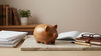Wooden piggy bank on desk with books, papers and glasses representing savings, financial planning and money management for education expenses.