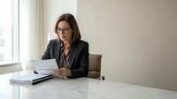Professional businesswoman in glasses reviewing documents at modern office desk with natural lighting for corporate presentations and workplace content.