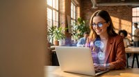 Young professional woman with glasses working on laptop in modern coworking office space with warm natural lighting and brick walls.
