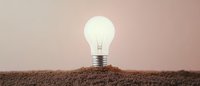 Glowing light bulb emerging from brown soil against neutral background symbolizes growth, innovation, and sustainable green energy solutions.