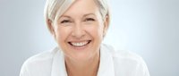 Portrait of a mature woman with short white hair, fair complexion, and a bright, genuine smile wearing a white shirt against a neutral gray background.