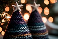 Knitted Christmas tree decorations with silver star toppers against blurred warm bokeh lights, creating cozy holiday atmosphere. Handmade festive ornaments.
