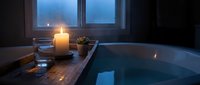 Serene bathroom spa setting with lit candle, succulent plant, and glass of water beside modern bathtub at dusk with window view.