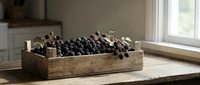 Young black grapes in rustic wooden crate with candles, displayed on table near window for harvest decoration and home styling.