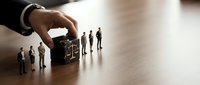 Business hand selecting candidate from group of miniature people figures on wooden desk representing recruitment and human resources management.