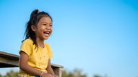 Young Asian girl wearing bright yellow shirt laughing outdoors against clear blue sky, expressing joy and happiness for lifestyle, childhood, and positive emotion concepts.