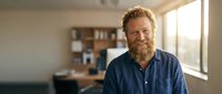 Smiling middle-aged Caucasian man with red beard in blue shirt standing in modern office workspace with natural lighting for business portraits.
