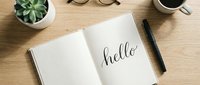 Open notebook with hello handwritten in calligraphy on wooden desk, pen, coffee cup, glasses, and succulent plant nearby.