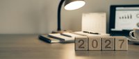 Wooden blocks spelling 2027 on a desk with lamp, notebook, and laptop in background, symbolizing new year business planning and goals.