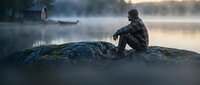 Young man sitting alone on mossy rock by misty lake at dawn, contemplating peaceful morning scenery with soft golden light and calm water reflections.