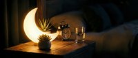 Cozy bedroom nightstand with glowing crescent moon lamp, alarm clock and glass of water creating warm ambient lighting for peaceful sleep atmosphere.