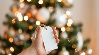 Caucasian hand holding blank white gift tag with string against blurred holiday lights and decorated tree, suitable for seasonal marketing and festive promotional campaigns.