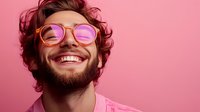Young bearded man wearing pink tinted glasses smiling broadly against pink background, showing genuine happiness and positive emotions. Modern lifestyle portrait.
