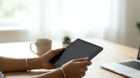 Cropped view of woman holding tablet device at wooden desk with coffee mug and laptop, bright window background, workspace setup for business, technology, and remote work concepts.