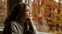 Thoughtful young woman with curly hair gazing through window at colorful autumn foliage, wearing cozy sweater in warm indoor setting for seasonal lifestyle content.