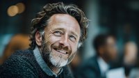 Happy mature Caucasian businessman with curly gray hair and beard smiling warmly in modern office environment during professional meeting or conference.