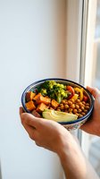 Hands holding colorful vegan Buddha bowl with roasted vegetables, chickpeas, broccoli and avocado for healthy plant-based nutrition and wellness lifestyle.