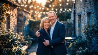 Happy mature Caucasian couple embracing outdoors with warm string lights creating romantic bokeh background for anniversary celebration.