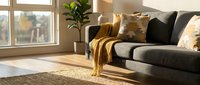 Modern living room with dark gray sofa, gold and black pillows, mustard throw blanket, potted plant, and natural sunlight from large window overlooking autumn landscape.