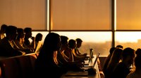 Business professionals working on laptops during golden hour meeting in modern office conference room with warm sunset lighting through windows.