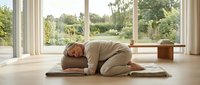 Middle-aged woman with light skin tone practicing child's pose yoga on mat in bright, minimalist living room with large windows overlooking garden. Wellness and relaxation.