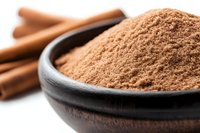 Ground cinnamon powder in rustic wooden bowl with whole cinnamon sticks in background, macro view showing aromatic spice texture and natural brown color.