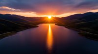 Dramatic sunset over mountain lake with golden sun rays reflecting on calm water surface, creating vibrant orange path amid silhouetted hills and dramatic purple clouds.