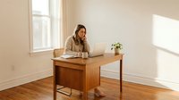 Young professional woman working on laptop at wooden desk in bright minimalist home office with natural sunlight streaming through windows.