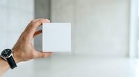 Male hand holding blank white card or paper against blurred interior background, suitable for mockups, branding, and advertising presentations.