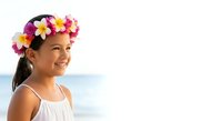 Young girl with long dark hair wearing white sleeveless dress and colorful flower crown with pink, yellow, and magenta blooms, smiling outdoors near beach.