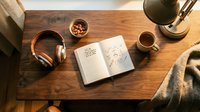 Cozy workspace flat lay with open notebook, headphones, coffee cup, and snacks on a wooden desk in warm ambient light.
