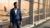 Professional African American businessman in navy suit standing confidently at airport terminal window with aircraft in background during golden hour.