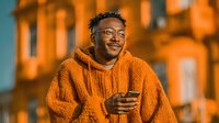 Happy young African American man in orange hoodie using smartphone outdoors at sunset with warm golden lighting and urban architecture background.