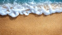 Gentle ocean wave with white foam washing onto golden sandy beach, creating natural border between sea and shore. Aerial perspective of coastline.