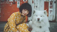 Young Asian woman in yellow floral dress sitting with fluffy white Samoyed dog against rustic door background, creating warm companionship moment.