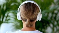 Young Caucasian woman with hair bun wearing white wireless headphones viewed from behind against green tropical foliage background, listening to music or podcast.