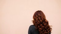 Woman with long wavy auburn hair viewed from behind against neutral beige background, suitable for hair care, beauty, and wellness marketing materials.