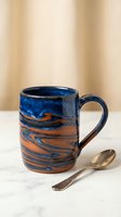 Handcrafted ceramic mug with blue and orange swirl pattern sits beside golden spoon on marble surface against warm beige curtain backdrop for kitchen decor.