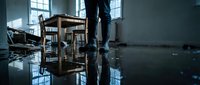 Man standing in flooded office space with overturned furniture and water damage reflecting natural light from windows creating dramatic indoor disaster scene.