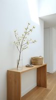 Minimalist wooden console table with flowering branch in glass vase and woven basket in modern white interior hallway for home decor styling.