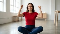 Young Hispanic woman in red shirt celebrating success with raised fists while sitting cross-legged on floor in bright modern apartment interior.