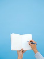 Person with medium skin tone holding open notebook with pen, writing or taking notes against blue background for business, education, planning, journaling, or creative work concepts.