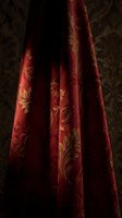Luxurious crimson velvet fabric with ornate gold floral damask pattern drapes elegantly against dark backdrop. Rich textile features intricate botanical motifs ideal for upholstery, interior design, fashion, and decorative projects.