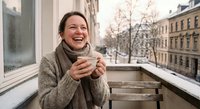 Happy young Caucasian woman laughing while holding warm drink on urban balcony during winter morning, enjoying cozy moment in knitted sweater outdoors.