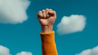 Raised fist in orange sweater against blue sky with white clouds symbolizing empowerment, protest, solidarity and determination for social justice movements.