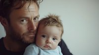 Caucasian father holding baby close, intimate family portrait showing emotional connection and paternal love. Soft natural lighting creates gentle mood and atmosphere.